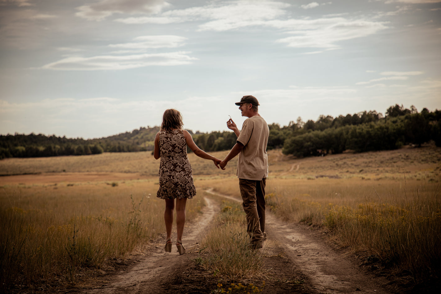 Two people walking hand in hand on a dirt path through a field.